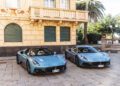 Two blue Maserati sports cars are parked side by side in front of a yellow building with closed green shutters in Italy, palm trees swaying behind—a perfect scene for an MCPURA Driving Experience.