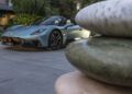 A blue Maserati sports car, inspired by the spirit of Italy, is parked on a paved driveway near stacked decorative stones and lush greenery.