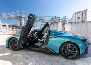 A teal Maserati sports car with its driver-side door open upward is parked outdoors on a concrete surface near industrial structures, reminiscent of an MCPURA Driving Experience in Italy.