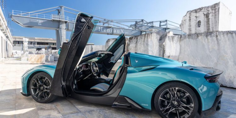 A teal Maserati sports car with its driver-side door open upward is parked outdoors on a concrete surface near industrial structures, reminiscent of an MCPURA Driving Experience in Italy.