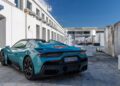 A teal Maserati sports car is parked on a stone surface outside a white industrial building under a clear blue sky, capturing the spirit of Italy and the thrill of an MCPURA Driving Experience.