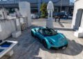 A turquoise Maserati convertible sports car is parked on a marble platform near a statue of an astronaut, with people and industrial buildings in the background—perfect for an unforgettable MCPURA Driving Experience in Italy.