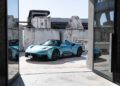 A turquoise Maserati MC20 sports car is parked in an industrial-style outdoor area with concrete walls and buildings in the background, promising an exhilarating driving experience.