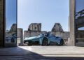 A blue Maserati sports car is parked on a concrete surface framed by open glass doors, with sunlit stone ruins in the background, evoking the spirit of Italy and the thrill of an MCPURA Driving Experience.