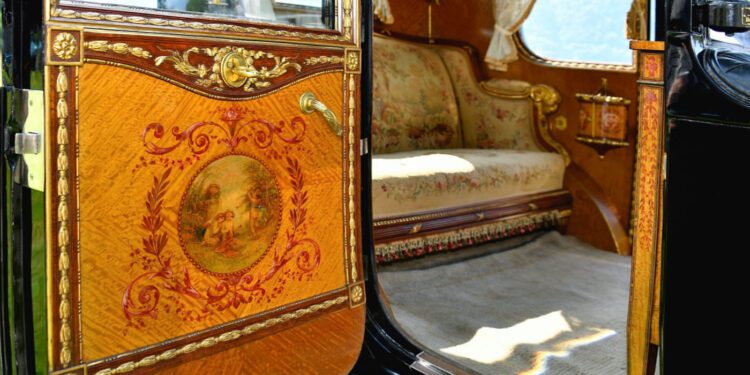 The interior of an ornate vintage carriage with elaborate woodwork, floral upholstery, and decorative painted details on the door evokes the bespoke luxury found in a classic Rolls-Royce Phantom.