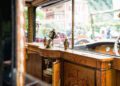 Interior of a vintage Bespoke Rolls-Royce featuring ornate wooden cabinetry, decorative vases, a clock, and glassware, with classic cars and greenery visible through the window—celebrating 100 Years of Rolls-Royce Phantoms.