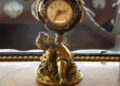 An ornate antique clock held by a gold cherub figurine sits on a decorative wooden surface, with a bespoke Rolls-Royce car interior visible in the background.