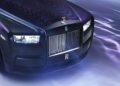 Close-up view of the front of a bespoke black Rolls-Royce car, featuring the signature grille, headlights, and hood ornament, with purple and blue light reflections on the surface.