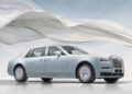 A light blue Rolls-Royce luxury sedan is parked on a white surface with flowing white fabric in the background under a clear sky.