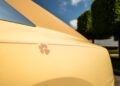 Close-up of a yellow Rolls-Royce Spectre with a red paw print decal on the side, parked outdoors near trimmed trees under a blue sky.