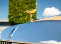 Close-up of the gold Spirit of Ecstasy and Rolls-Royce emblem on a bespoke Rolls-Royce, with a blurred tree and sky in the background.