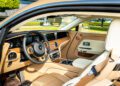 The interior of this Bespoke Rolls-Royce features tan leather seats, wood trim, a multi-function steering wheel, and a digital dashboard with various controls.