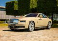 A beige Bespoke Rolls-Royce coupe, possibly a Rolls-Royce Spectre, is parked on a cobblestone area with manicured trees and a modern building in the background.