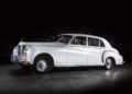 A classic bespoke white Rolls-Royce sedan is parked indoors under low lighting, highlighting its elegant side and front details.