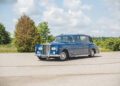 A classic blue Rolls-Royce sedan, part of 100 Years of Bespoke Phantoms, is parked on a paved surface with trees and a cloudy sky in the background.
