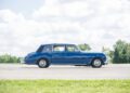 A classic blue Rolls-Royce sedan with chrome details is parked on an empty pavement, framed by trees and clouds in the background.