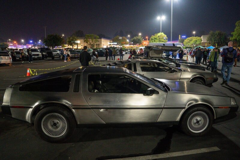 A DeLorean car is parked at a nighttime car meet in a parking lot for Film Friday, with more DeLoreans and people in the background under artificial lights.