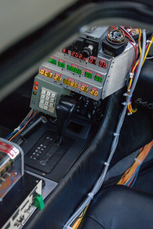 A car interior featuring a retro-styled electronic control panel with digital displays, buttons, and wiring, mounted beside the gear shift—perfect for a true Outatime moment.
