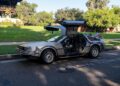 A silver DeLorean car with gull-wing doors open is parked on a street beside a grassy area, its "OUTATIME" license plate catching the eye—perfect for a Film Friday feature. Trees and buildings form the backdrop.