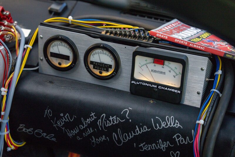 A close-up of a car dashboard with three meters, colorful wires, a "Plutonium Chamber" label, sports almanac, autographs from Claudia Wells and Bob Gale—pure Outatime nostalgia perfect for any Film Friday feature.