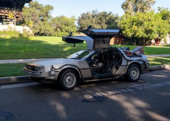 A silver DeLorean car with gull-wing doors open is parked on a street beside a grassy area, its "OUTATIME" license plate catching the eye—perfect for a Film Friday feature. Trees and buildings form the backdrop.