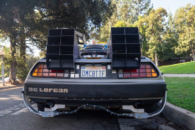 Rear view of a modified DeLorean with an "Outatime" vibe, license plate "DMCBTTF," parked on a street surrounded by trees and grass—a true Petrolicious moment.
