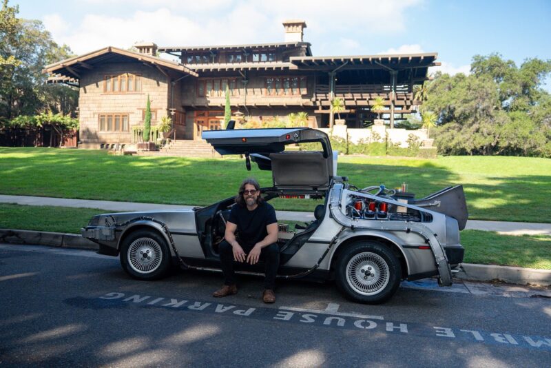 A man sits in a DeLorean with gull-wing doors open, parked in front of a large Craftsman-style house on a sunny day—a perfect Outatime moment for Petrolicious Film Friday.