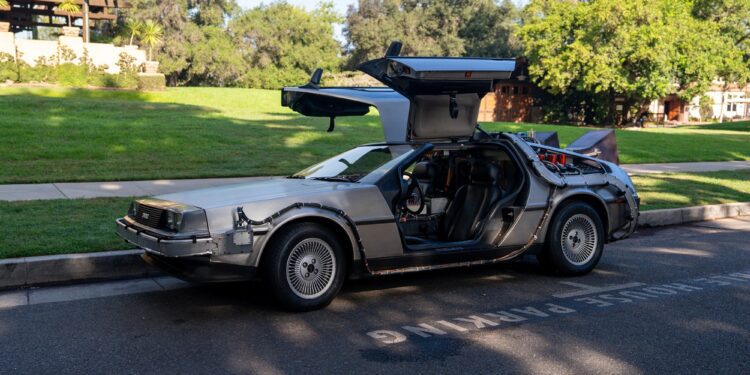 A silver DeLorean car with gull-wing doors open is parked on a street beside a grassy area, its "OUTATIME" license plate catching the eye—perfect for a Film Friday feature. Trees and buildings form the backdrop.