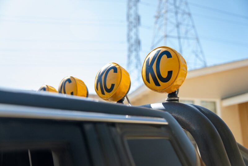 A row of yellow off-road lights with "KC" covers is mounted on top of a black vehicle, giving off serious Petrolicious vibes, with buildings and power lines visible in the background.