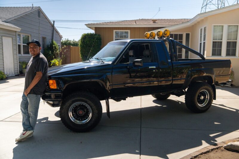 A person stands next to a black pickup truck with roll bar and roof lights, parked in a sunny residential driveway—perfect for a Petrolicious Film Friday feature.