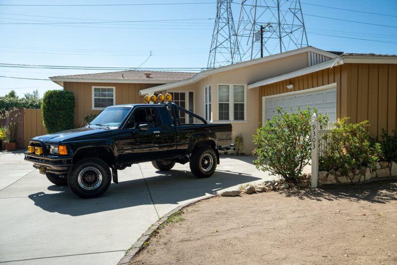 A black pickup truck with an "Outatime" vibe is parked in the driveway of a single-story house with tan siding and a white garage door.