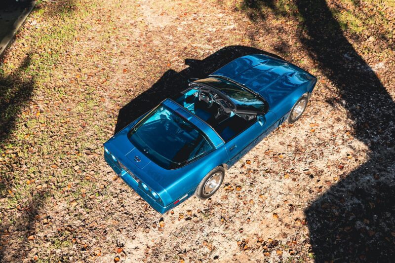 An overhead view of a blue Callaway Corvette convertible sports car parked on dry grass and fallen leaves, with sunlight casting shadows—a true Petrolicious moment.