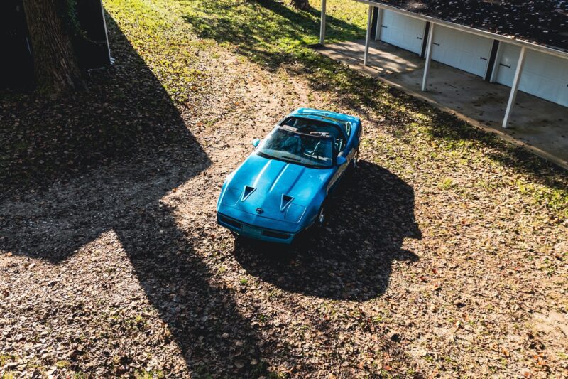 A blue Callaway Corvette with a targa top is parked on a leaf-covered dirt path beside a building with white garage doors, as if ready for its own Petrolicious Film moment.