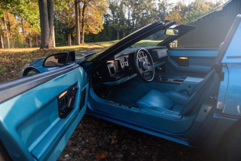 A blue Callaway Corvette convertible with the driver's door open reveals a matching blue interior and dashboard, parked outdoors on a leaf-covered ground near trees—a scene perfect for Petrolicious Film Friday.