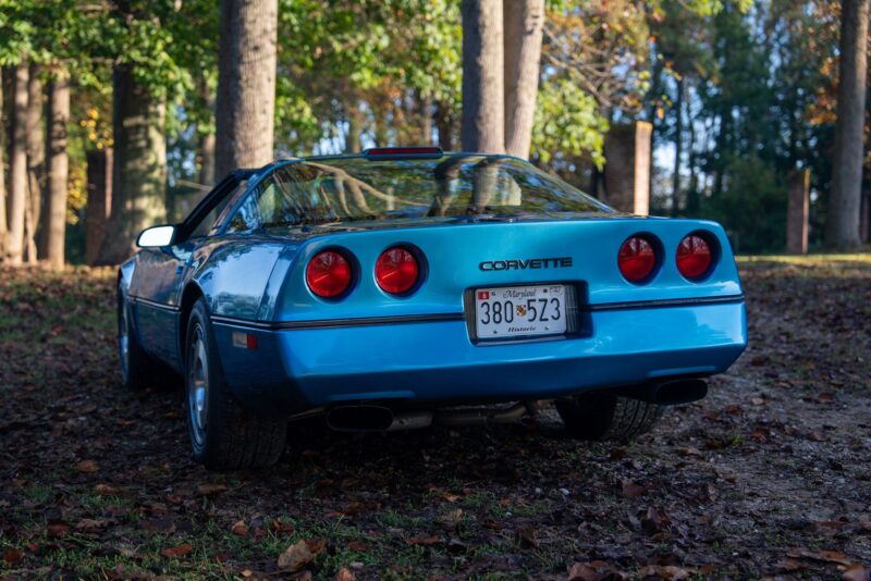 A blue Callaway Corvette is parked outdoors on a leaf-covered ground, viewed from the rear with trees and sunlight in the background—a perfect scene for a Petrolicious Film Friday.