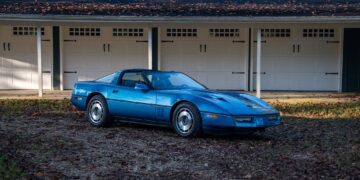 A blue Callaway Corvette, worthy of a Petrolicious Film Friday feature, is parked on a gravel surface in front of a row of white garage doors.