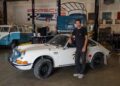 A man stands next to a vintage white Porsche sports car in a garage, with a blue Volkswagen van and various automotive tools in the background.