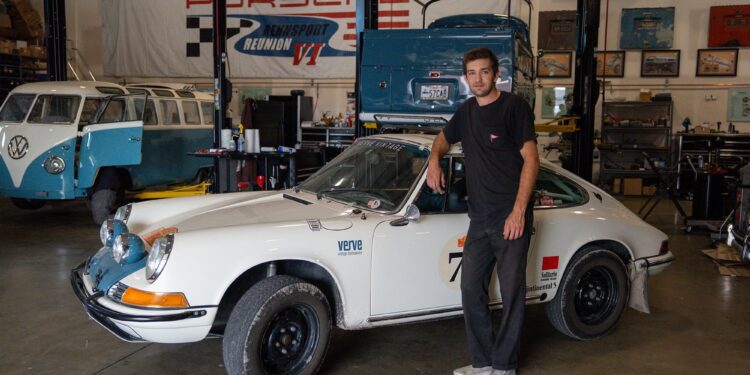 A man stands next to a vintage white Porsche sports car in a garage, with a blue Volkswagen van and various automotive tools in the background.