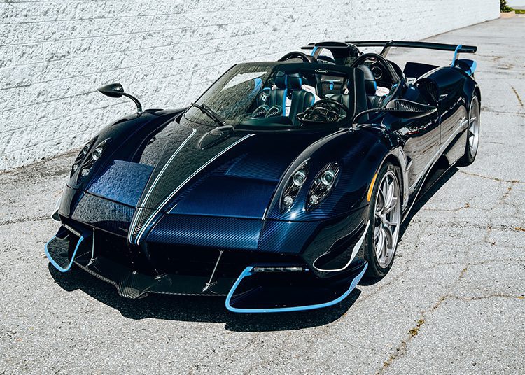 A black and blue Pagani Huayra Roadster, boasting 864HP, is parked on cracked pavement near a white brick wall.