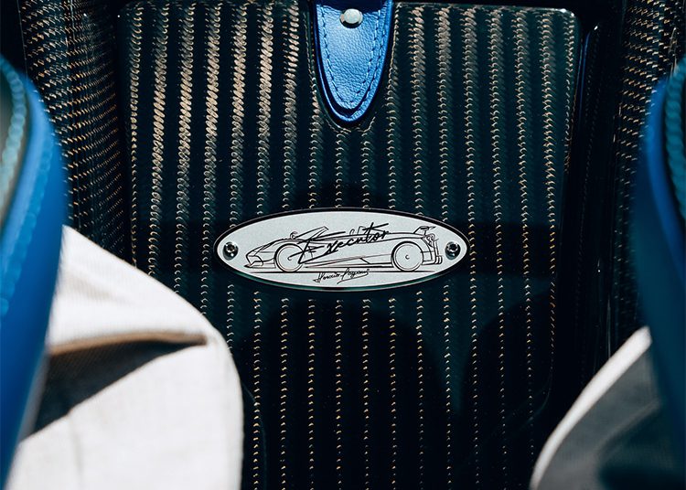 Close-up of a metal Bugatti nameplate mounted on a carbon fiber panel, surrounded by blue and beige materials—a detail captured at O'Gara Coach, specialists in luxury brands like Pagani Huayra Roadster.