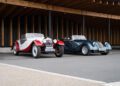 Two classic convertibles—a Morgan Plus Four in white and red and a dark blue roadster—are parked side by side under a modern, wooden canopy, celebrating the Morgan 75th Anniversary in style.