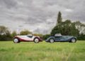 Two vintage-style convertibles—a red and white Morgan Plus Four and a dark green classic—are parked side by side on a grassy field beneath a cloudy sky, celebrating the 75th Anniversary of iconic automotive craftsmanship.