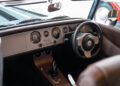 Interior view of a Morgan Plus Four Anniversary shows the dashboard, steering wheel, analog gauges, and automatic gear shifter with elegant wood paneling on the center console.