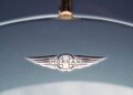 Close-up of the Morgan car logo with silver wings and "Morgan" text on the hood of a blue Morgan Plus Four, celebrating the brand's 75th Anniversary.