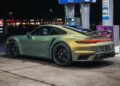A green Porsche 911 sports car is parked at a gas station pump at night, illuminated by the lights from the station and nearby buildings—an eye-catching scene for those searching for exclusive Porsches or browsing Porsches for sale.