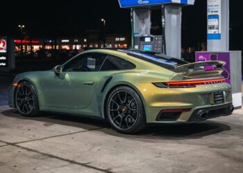 A green Porsche 911 sports car is parked at a gas station pump at night, illuminated by the lights from the station and nearby buildings—an eye-catching scene for those searching for exclusive Porsches or browsing Porsches for sale.