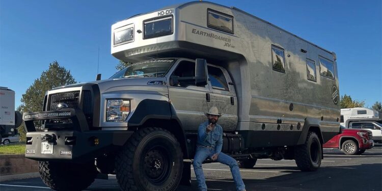 A person in a hat and jeans sits on the front bumper of a large off-road EarthRoamer HD RV parked in a lot on a clear day.