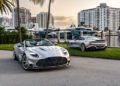 Two silver convertible sports cars are parked near a marina with yachts and high-rise buildings in the background.