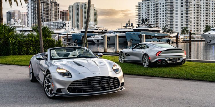 Two silver convertible sports cars are parked near a marina with yachts and high-rise buildings in the background.