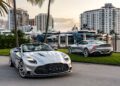 Two silver luxury convertibles parked near palm trees and yachts at a marina, with high-rise buildings in the background.